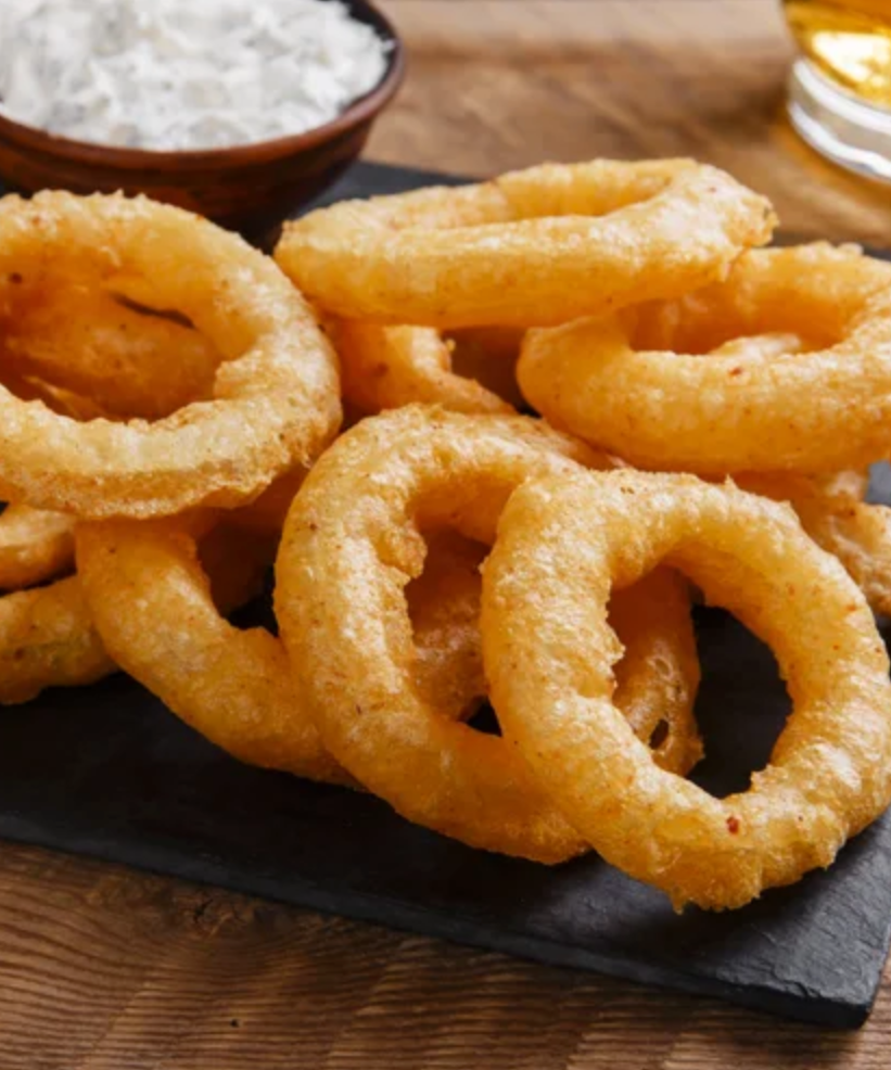 Battered onion rings on a black slate with a glass of beer and a bowl of coleslaw in the background.
