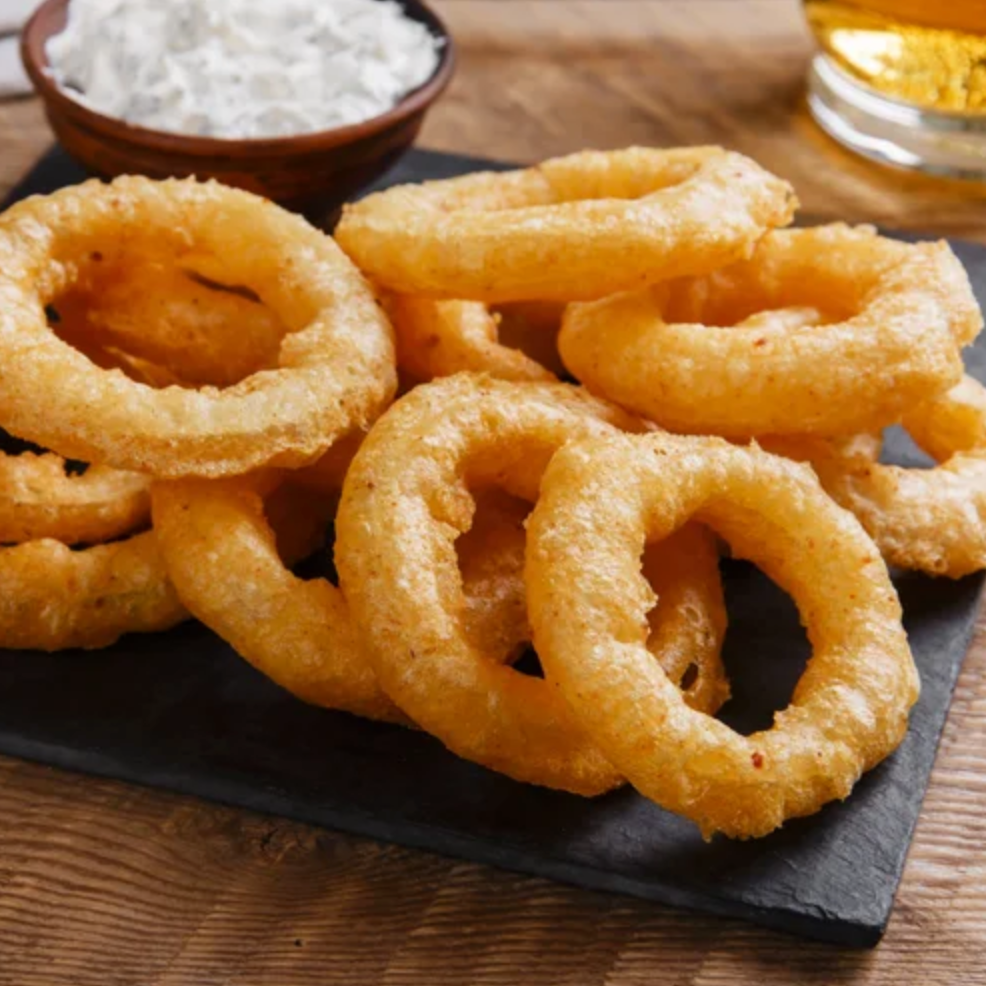 Battered onion rings on a black slate with a glass of beer and a bowl of coleslaw in the background.